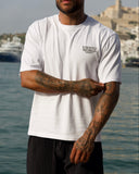 A man wearing a white crew-neck t-shirt with a small black logo on the left chest and black pleated shorts, standing in front of a scenic coastal background with water and buildings.