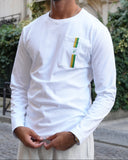 A man wearing a white long-sleeve pocket shirt with a small green and yellow striped accent on the chest pocket, paired with light gray sweatpants, standing on a cobblestone street in front of a classic stone building.