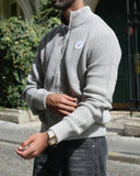 A man wearing a light gray waffle-knit mock neck sweater and dark gray jeans, standing on a cobblestone street in front of a stone building.