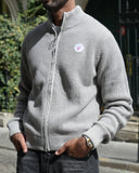 A man wearing a light gray waffle-knit zip-up sweater and dark gray jeans, standing on a cobblestone street in front of a historic building.