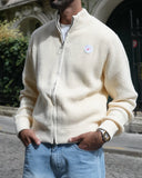 A man wearing a cream-colored waffle-knit zip-up sweater and light blue jeans, standing on a cobblestone street in front of a classic stone building.