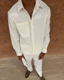 A man wearing a white long-sleeve button-up shirt with a chest pocket, paired with white trousers and brown slip-on shoes, standing against a textured brown wall.