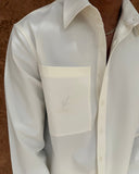 A close-up of a person wearing a white long-sleeve button-up shirt with a large chest pocket featuring a subtle embroidered logo, set against a textured brown background.