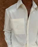 A close-up of a person wearing a white long-sleeve button-up shirt with a large chest pocket and a subtle embroidered logo, set against a textured brown background.