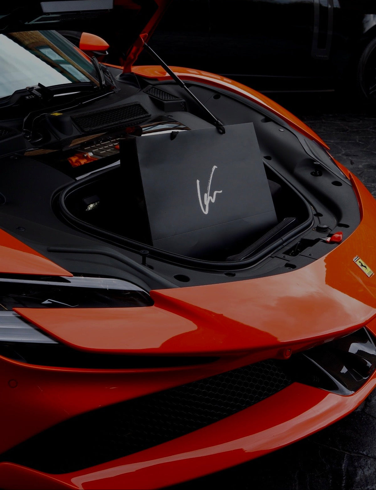 An orange sports car with its hood open, showing a black shopping bag with a white script logo inside the trunk. A black SUV is parked in the background near a brick building.